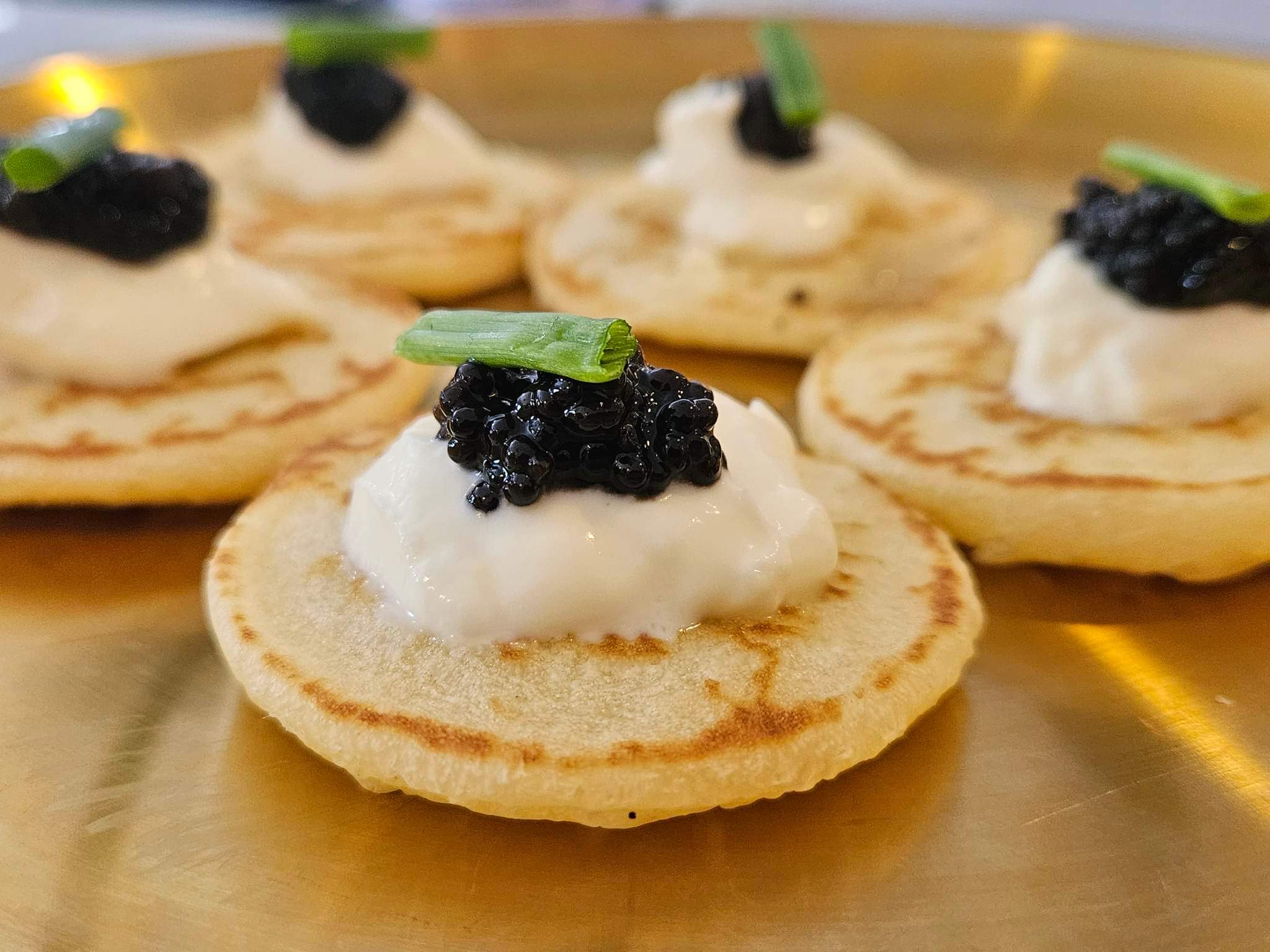 caviar appetizer constisting of 5 french belinis toped with a creme fraiche caviar and finished with chive plated on a gold plate