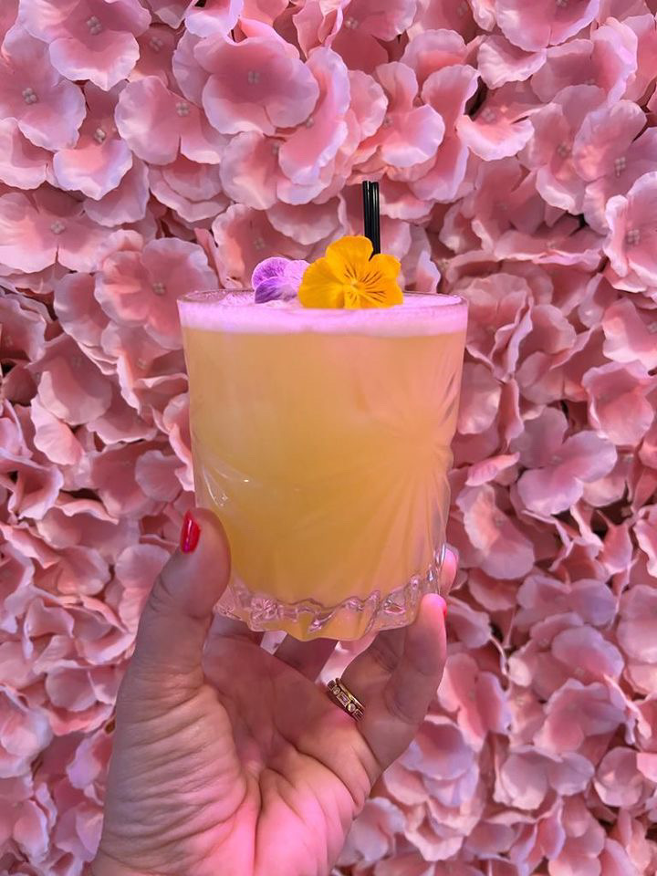 a hand holding a paloma drink topped with edible flowers with a flower wall background